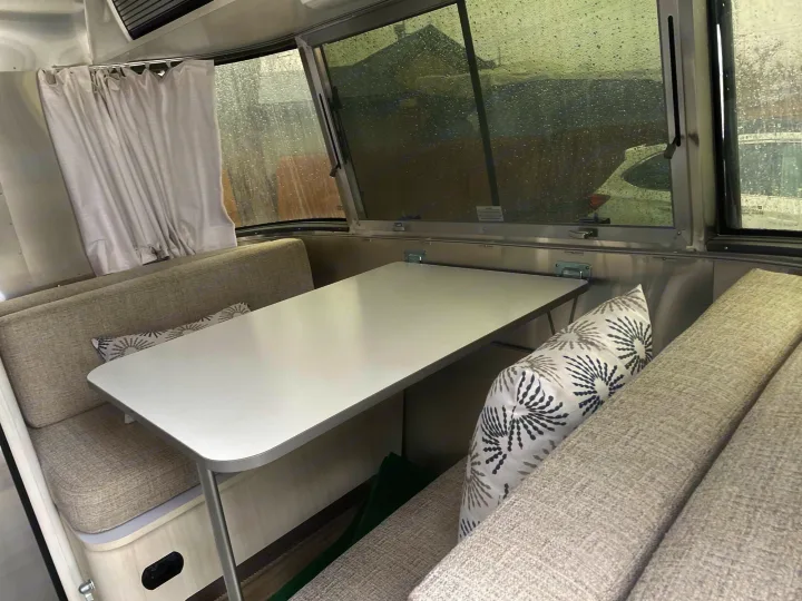 Enjoy this clean RV rental interior with dining table and cozy benches, perfect for any weather. Rain visible outside.. Airstream Bambi 2021