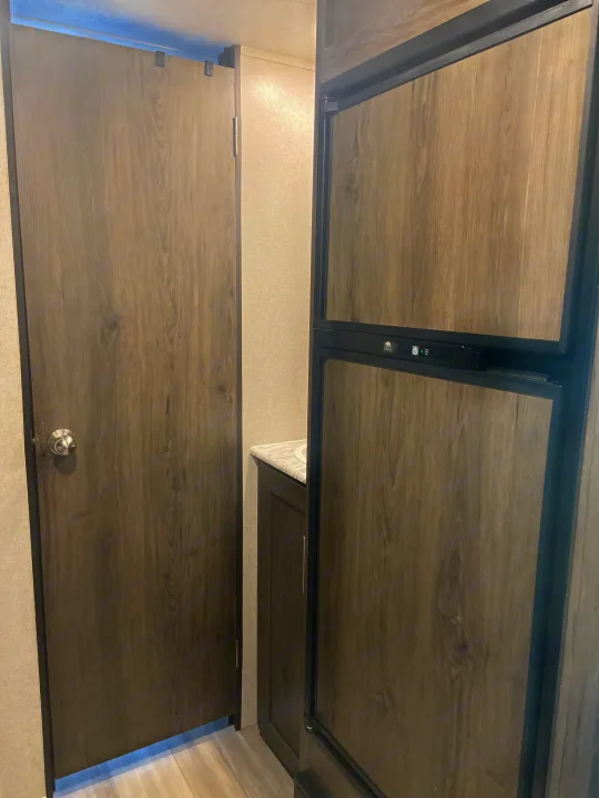 An RV rental interior with a wood-look refrigerator and a wooden door, hinting at a compact bathroom beyond.. Clipper Other 2021