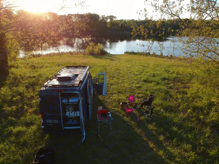 An RV rental camper van parked by a serene lake at sunset, with a person relaxing in a chair, ideal for camping.. vandoit DO 2018