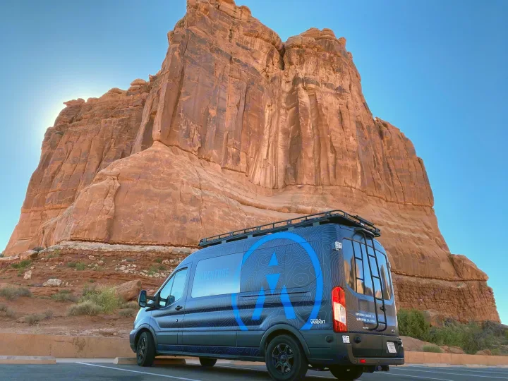This dark blue RV rental van, with roof rack & custom graphics, is parked against a backdrop of towering red rock cliffs.. vandoit DO 2018
