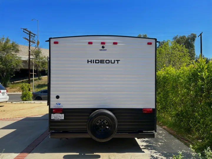 Rear view of a white and black Hideout travel trailer with a spare tire, ready for RV rental. Parked on a sunny driveway for outdoor trips.. Keystone RV Hideout 2022
