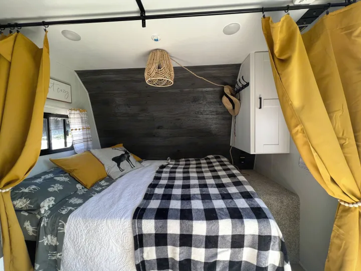 An RV rental's cozy bedroom features a bed with plaid and deer pillows, yellow curtains, and wood paneling.. Keystone RV Hideout 2022