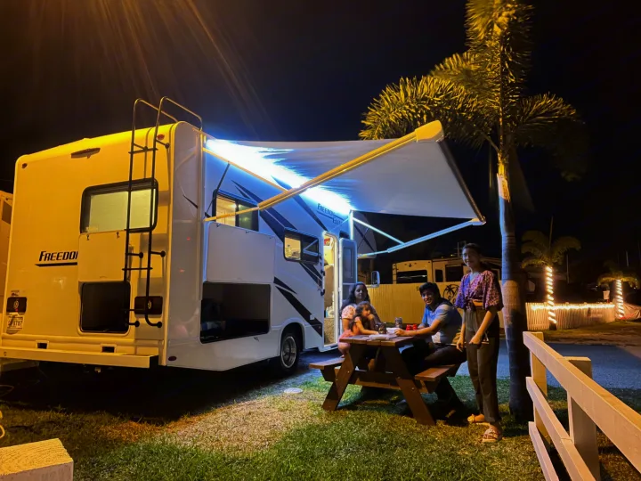 A family enjoys their RV rental with bright awning lights at night. They're gathered around a picnic table in a scenic campsite.. Thor Freedom Elite 2021