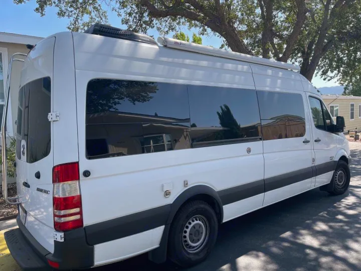 A white Mercedes Sprinter camper van, ideal for rv rental, is parked outdoors. Features tinted windows, rooftop AC, and side awning.. Sportsmobile Custom Design 2013