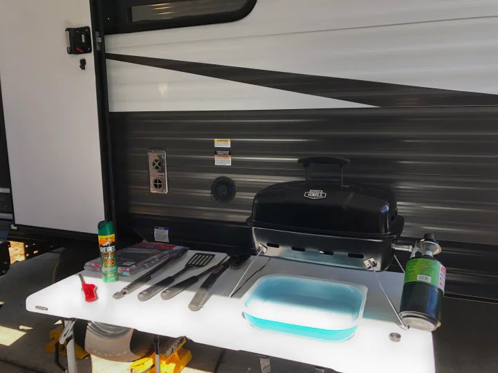 An RV rental features an outdoor grilling station with a portable grill, propane, and utensils on a white table beside the camper.. Forest River Ozark 1800 QS 2020