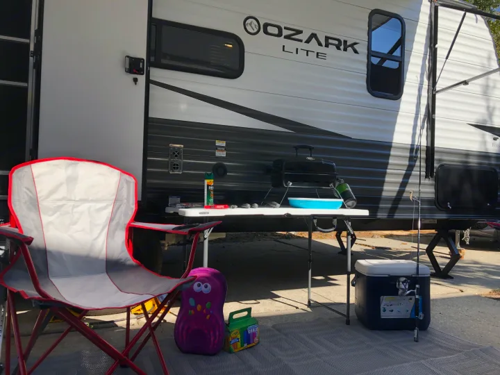 Relaxing at an RV rental campsite: chair, grill, cooler, fishing rod & Ozark Lite travel trailer setup.. Forest River Ozark 1800 QS 2020