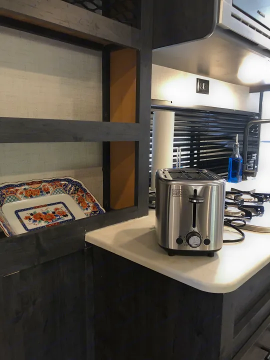 This RV rental kitchen features a sleek Black+Decker toaster, gas cooktop, and stylish wall decor, ready for use.. Forest River Ozark 1800 QS 2020