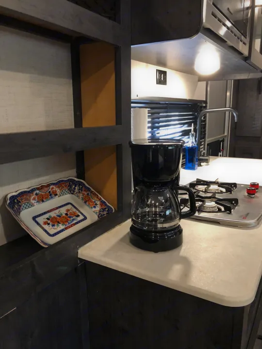 RV rental kitchenette showing coffee maker, 2-burner stovetop, and shelving with dish. Compact & functional interior.. Forest River Ozark 1800 QS 2020