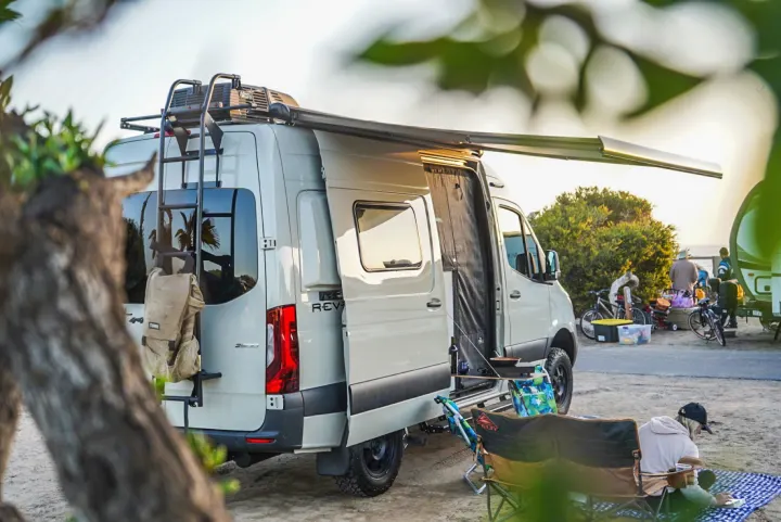 This rv rental shows a light camper van with an open side, extended awning, and outdoor cooking area at a lively campsite.. Winnebago Revel 2022