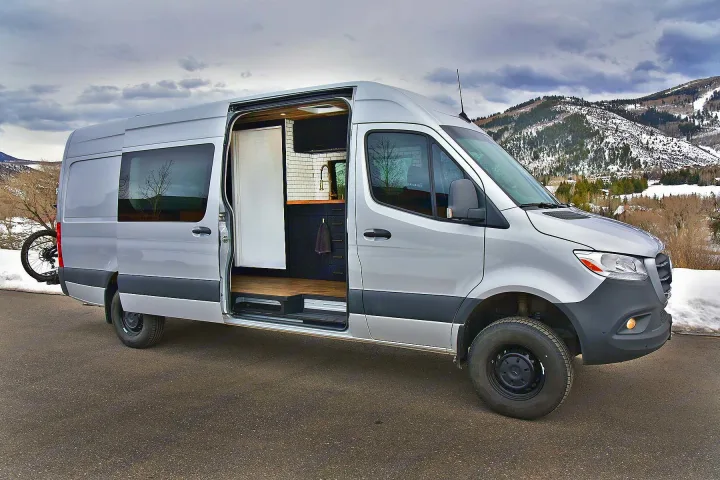 Silver Mercedes Sprinter rv rental camper van with its side door open, revealing a custom kitchen inside, parked against snowy mountains.. Mercedes-Benz Sprinter 2021