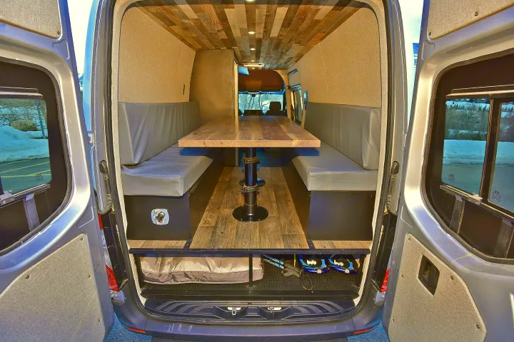 Spacious camper van interior for RV rental. Features a dining area, comfy benches, wood ceiling, and rear storage.. Mercedes-Benz Sprinter 2021