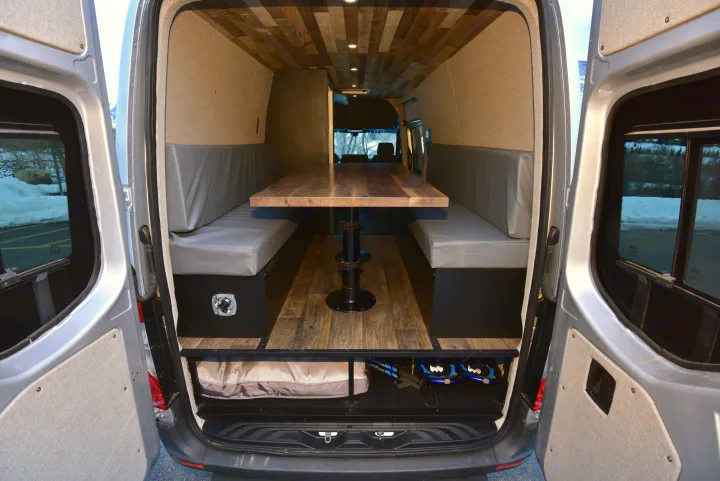 Inside an RV rental: a stylish camper van conversion with a wooden table, cozy benches, and ample rear storage.. Mercedes-Benz Sprinter 2021