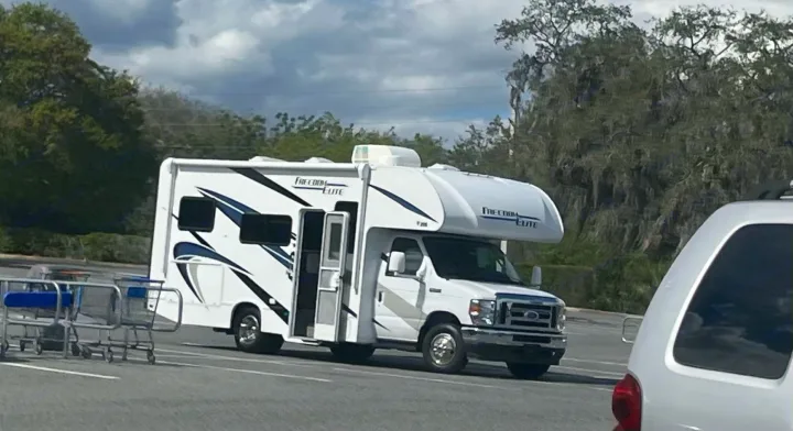 A large white "Freedom Elite" RV rental, door open, is parked in a sunny lot with trees and shopping carts nearby.. Thor Freedom Elite 2021
