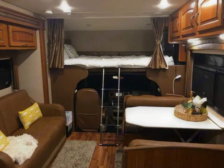 Cozy RV rental interior shows a sleeping bunk over the cab, living area, & dinette with a welcome basket.. Jayco Greyhawk 2016