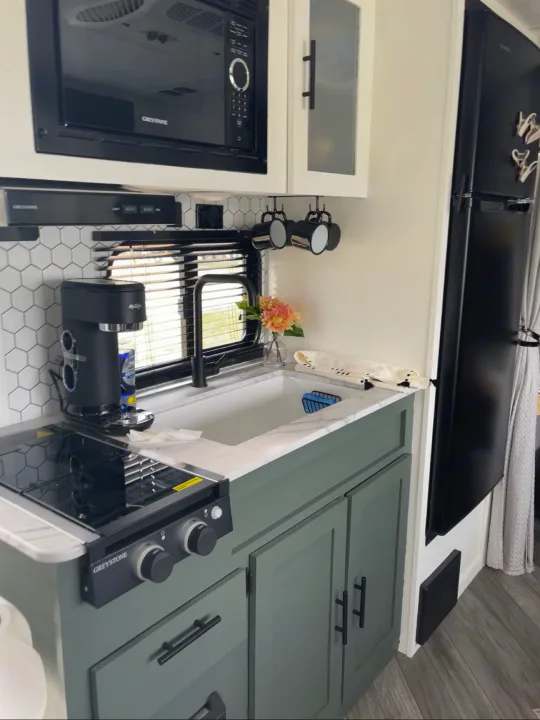 Modern RV rental kitchen with black appliances, including a microwave, stove, and fridge, light counters, and green cabinets.. Forest River 178BHSKX 2022