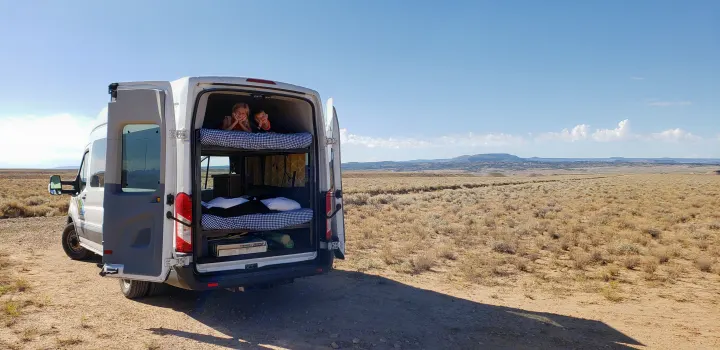 An RV rental camper van with open back doors reveals kids smiling from bunk beds in a vast, sunny desert landscape.. Ford transit 2017
