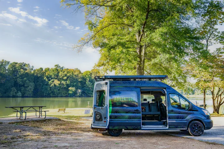 An Adventure Rig rv rental van, patterned in blue/green, is parked lakeside with its side door open, inviting outdoor travel.. Ford Transit Custom 2017