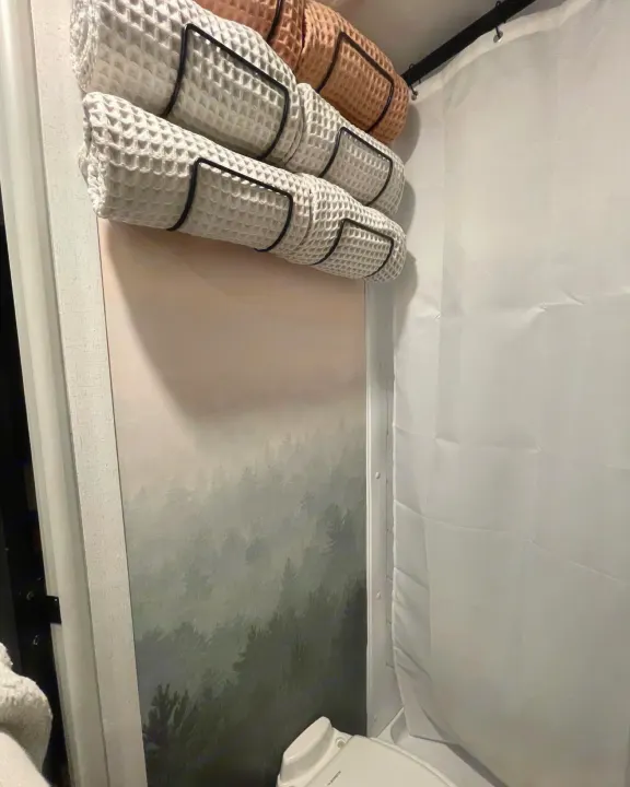 Compact RV rental bathroom featuring stylish rolled towel storage, a misty forest mural, and a white shower area.. Forest River 178BHSKX 2022
