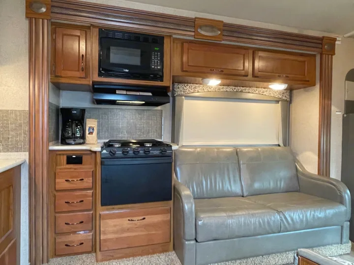 RV rental interior features a functional kitchen with wood cabinets, microwave, stovetop, and a comfortable light gray sofa.. Jayco Redhawk 2016