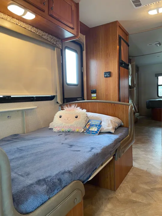 Bright RV rental interior: a child's bed with a llama plushie & books, facing a kitchen area and rear bunk.. Jayco Redhawk 2016