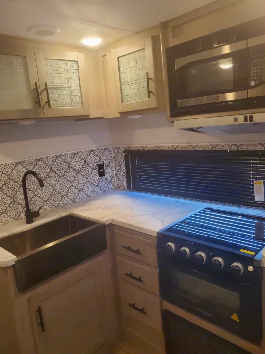 Modern RV rental kitchen showcasing a large sink, patterned backsplash, oven, microwave, and light-toned cabinets.. Coleman 2455bh 2022