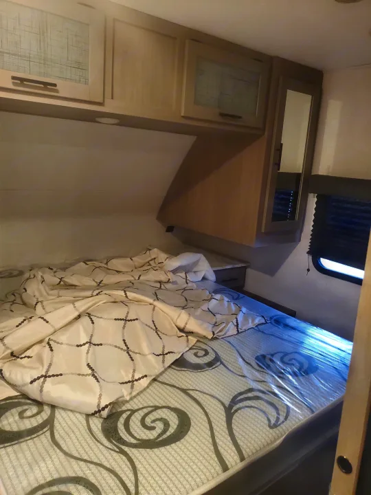 Inside an RV rental bedroom: a bed with patterned mattress and white comforter, overhead cabinets, and a side mirror.. Coleman 2455bh 2022