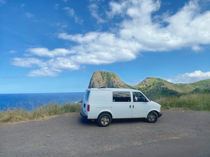 White RV rental van parked on a scenic coastal road, overlooking a blue ocean & green hills. A dog enjoys the view from inside.. Chevrolet Other 2005