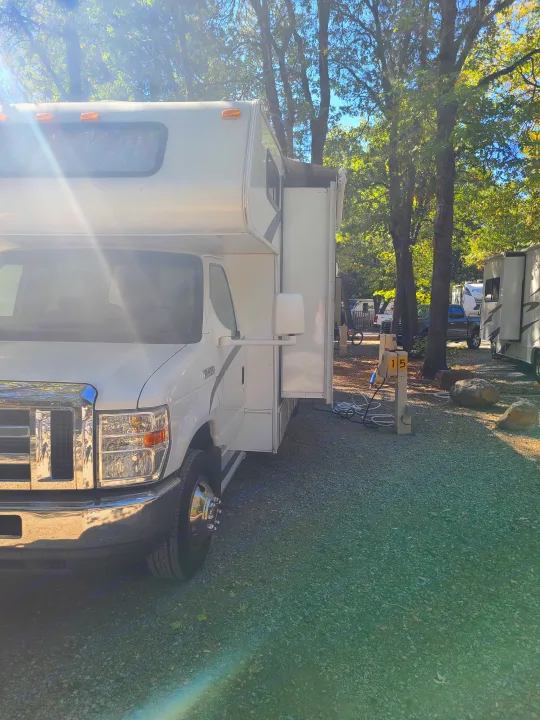 Sun-drenched white rv rental parked at campsite 15, slide-out extended, hooked up to power in a treed campground.. Coachmen Freelander 2011