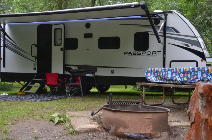 A white travel trailer RV rental at a green campsite with a blue-lit awning, picnic table, fire pit, and grill.. Keystone Passport 2021
