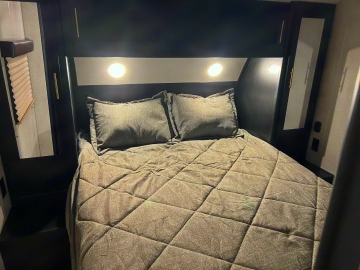 This RV rental bedroom features a cozy grey quilted bed, two pillows, overhead lights, and sleek dark storage.. Forest River Cherokee Grey Wolf 2022