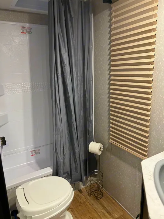 This RV rental bathroom features a compact white toilet, shower with gray curtain, pleated window blind, and TP holder.. Forest River Cherokee Grey Wolf 2022