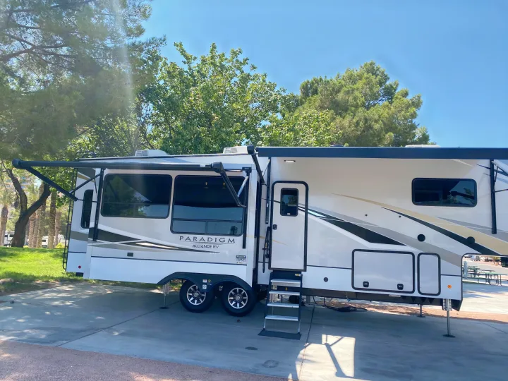 Experience outdoor comfort! This Paradigm Alliance RV rental, a white fifth wheel, is set up with awnings extended at a sunny camp.. Alliance RV Paradigm 2021