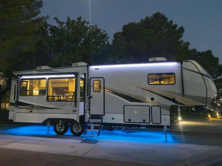 Luxury rv rental: A modern fifth-wheel RV glows with white awning lights, blue underglow, & warm interior lights at dusk.. Alliance RV Paradigm 2021