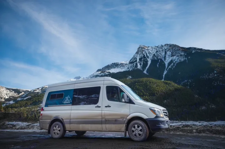 Adventure-ready RV rental: A beige Mercedes Sprinter campervan parked on a dirt road with snow-capped mountains and blue sky.. Mercedes Sprinter 2024