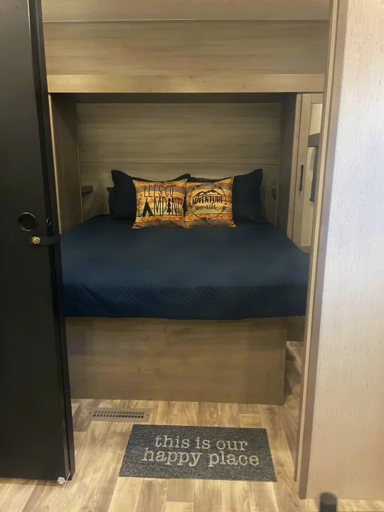 View of a compact RV rental bedroom with a dark blue bed, two camping-themed pillows, and a "happy place" floor mat.. Dutchman Kodiak SE 2022