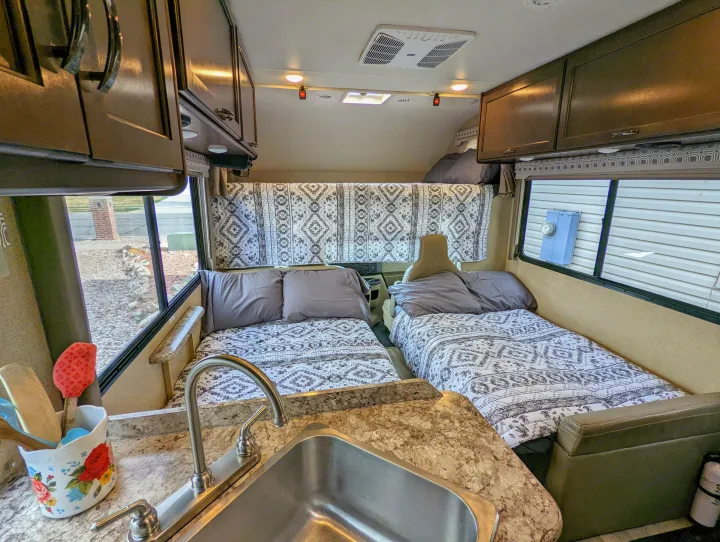 Inside a comfortable **RV rental**, showing two patterned beds, an equipped kitchen with a sink, and overhead cabinets for storage.. Thor Four Winds 2020
