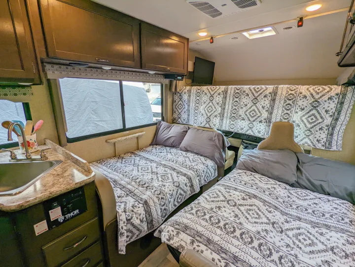 Inviting rv rental interior features two neatly made beds with patterned quilts, an equipped kitchen counter, & overhead cabinets.. Thor Four Winds 2020