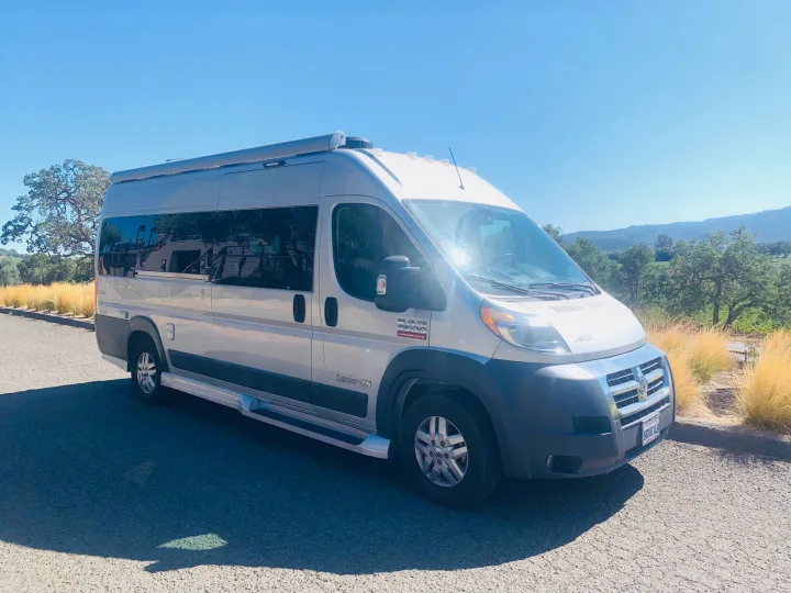 This silver van-style RV rental is parked on a paved road, bathed in sunlight against a backdrop of green hills and golden grass.. Pleasure Way Lexor 2017