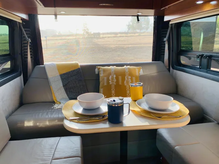 Bright RV rental interior: a cozy dining area set for two with white bowls, yellow placemats, and large scenic windows.. Pleasure Way Lexor 2017