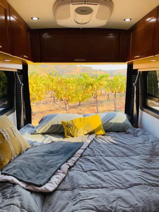 Inside an RV rental, a comfy bed faces a vibrant autumn vineyard. Relax & enjoy the scenic view!. Pleasure Way Lexor 2017