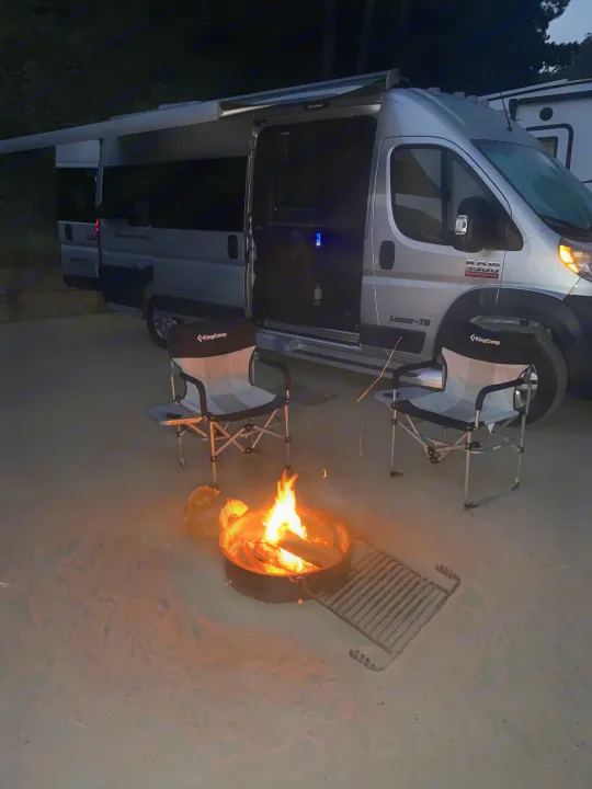 Cozy RV rental campsite at night: a silver campervan, two chairs, and a warm, glowing campfire on sandy ground.. Pleasure Way Lexor 2017