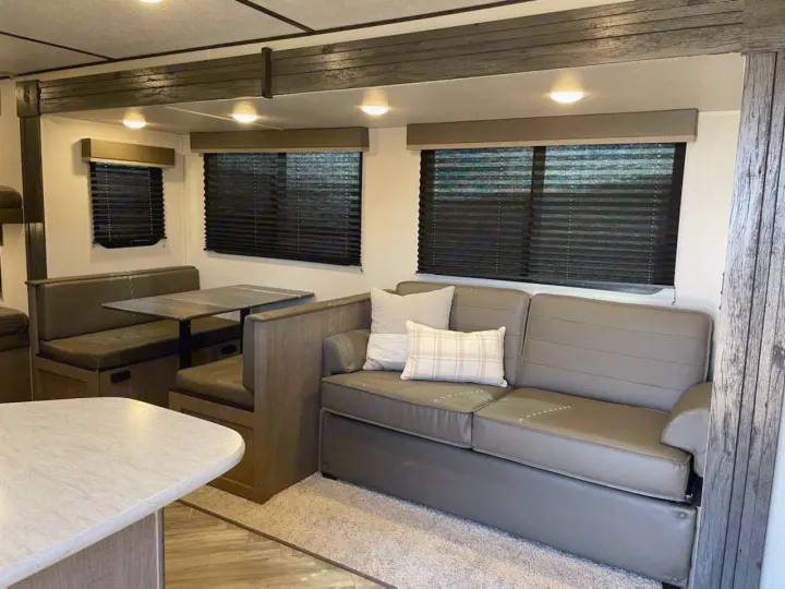 Spacious RV rental interior with a dining booth, a gray sofa with pillows, and shaded windows for comfortable travel.. Keystone RV Hideout 2021
