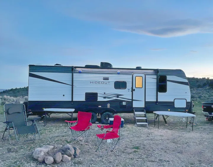 An RV rental Hideout travel trailer at a campsite, with illuminated awning lights, chairs, tables, and a rock campfire ring.. Keystone RV Hideout 2021