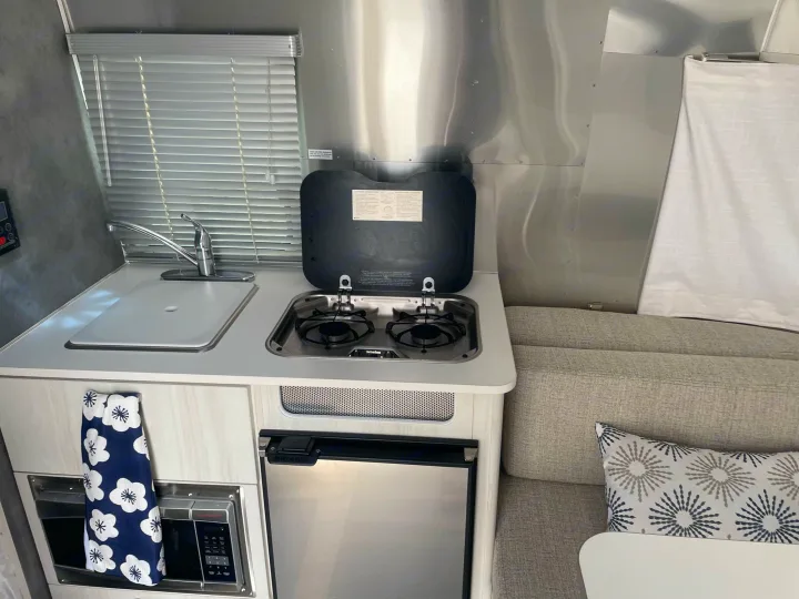 View of an RV rental kitchen showing a compact sink, stove, microwave, and small fridge, with cozy seating adjacent.. Airstream Bambi 2021