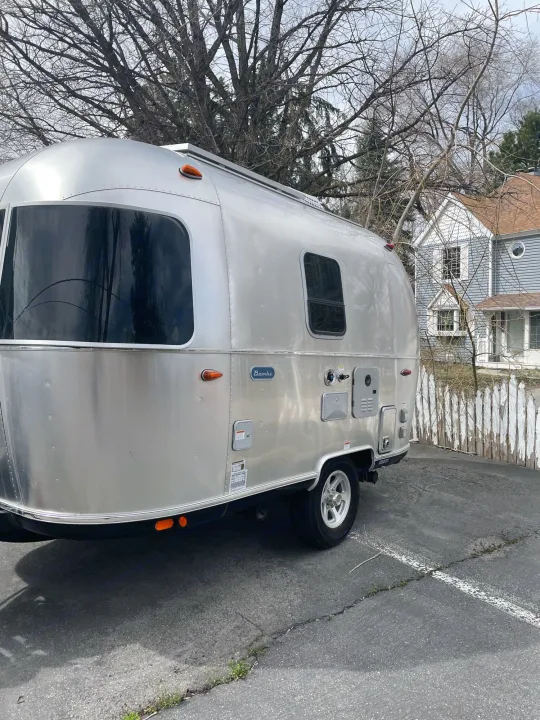 Shiny Airstream Bambi travel trailer. Consider this unique RV rental for your next trip, parked on asphalt by a fence.. Airstream Bambi 2021