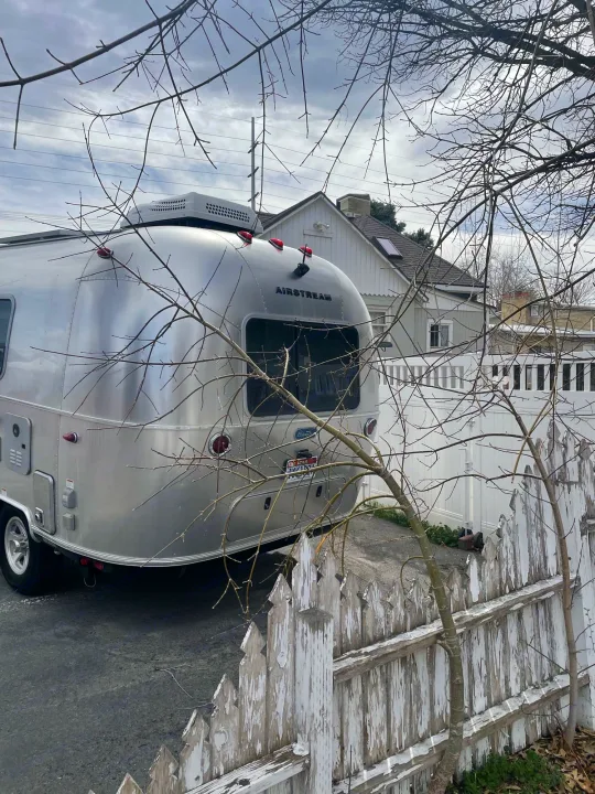 Shiny Airstream RV parked, ready for an rv rental adventure, behind a worn picket fence and leafless tree.. Airstream Bambi 2021