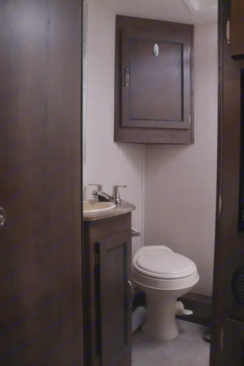 Compact RV rental bathroom interior with a toilet, sink, vanity, and overhead dark wood cabinet against textured walls.. Coachmen Freelander 2020