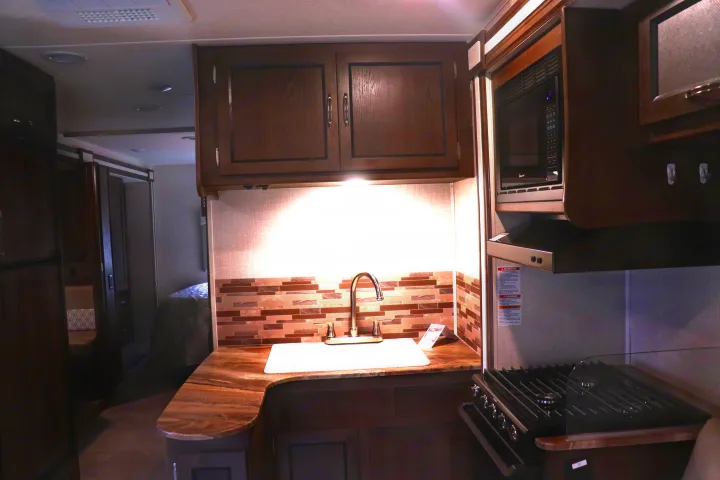 Modern RV rental kitchen with wood cabinets, sink, microwave, and stovetop. Bedroom visible in background.. Coachmen Freelander 2020