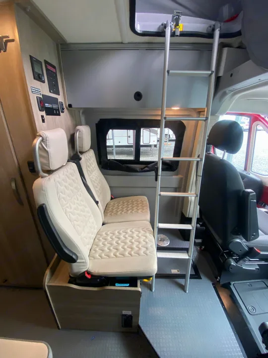 Interior view of an RV rental showing beige passenger seats, ladder to an overhead bunk, and wall-mounted controls.. Winnebago Solis 2022