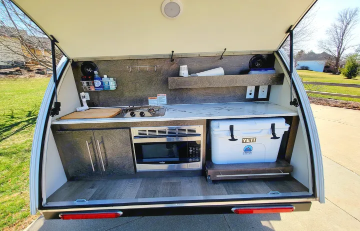 Explore this RV rental's compact outdoor kitchen galley in a teardrop camper, featuring a sink, stove, microwave, and YETI cooler.. nuCamp T@G 2020